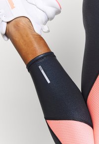 Leggings sportivi neri in tessuto strutturato, con un accento in rete rosa e una patch bianca con logo sul polpaccio. Progettati per l'abbigliamento attivo.