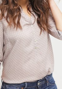 Camicia button-up di un leggero taupe con un motivo floreale, dotata di un colletto e cinque bottoni, indossata con jeans blu scuri.