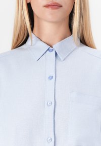 Světle modrá košile s knoflíky, vyrobená z texturované látky, s límcem typu point, šesti knoflíky a jednou náprsní kapsou.