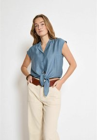 Camicetta blu a maniche corte con nodo frontale, realizzata in tessuto liscio. Abbinata a pantaloni beige a vita alta e una cintura marrone.