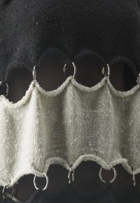 Tissu en maille noire avec un panneau en maille ondulée crème, relié par des anneaux argentés, présentant des textures contrastées et des bords en forme d'ondulations.