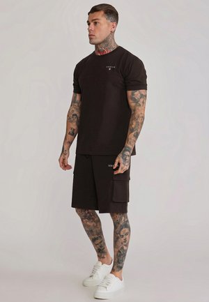 Muž s celotelovými tetovaniami nosí čierne tričko SikSilk a šortky s bielymi teniskami, stojí na jednoduchom pozadí.
