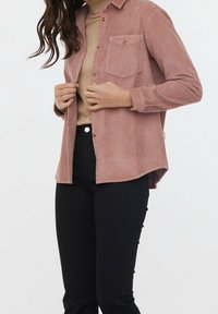 Femme portant une chemise en velours côtelé rose poussiéreux sur un col roulé beige, associée à un pantalon noir taille haute, debout devant un fond uni.