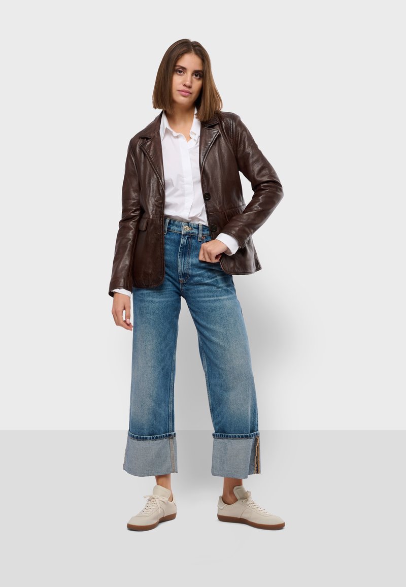 Giacca di pelle marrone con colletto, camicia bianca sotto, jeans blu a gamba larga con orli sfilacciati e sneakers color crema.