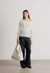 Biely pletený top s odhalenými ramenami, priehľadná vrstva, doplnený o tmavé rifle s vysokým pásom a veľkú krémovú kabelku typu hobo. Jemný, minimalistický dizajn.