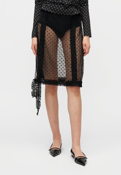 Jupe transparente à pois noirs avec des détails à volants, dotée d'un ourlet à franges. Associée à des ballerines noires pointues.