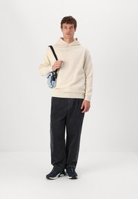 Sudadera beige, ajuste relajado, con capucha ajustable, combinada con pantalones de corduroy negros holgados. Un bolso cruzado azul y zapatillas oscuras completaron el look.