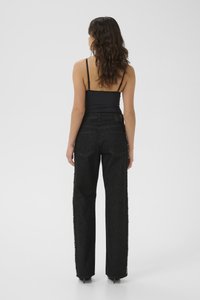 Mujer con cabello largo y ondulado viste una blusa negra sin mangas y jeans de talle alto de pierna ancha negros con adornos laterales, de pie de espaldas.