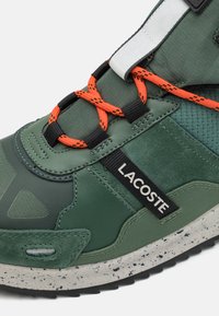 Zielone sneakersy z teksturowaną skórzaną i materiałową cholewką, wyposażone w czarne i pomarańczowe sznurówki, metkę z logo oraz nakrapianą gumową podeszwę.