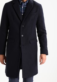 Abrigo de lana en un tono azul marino oscuro con un diseño estructurado, solapas de muesca, dos bolsillos frontales y cierre de botones, usado sobre una camisa a cuadros.