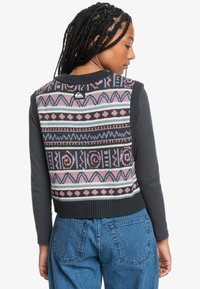 Chaleco de punto con patrones geométricos multicolores en rosa, azul y negro; dobladillo acanalado; etiqueta visible; superpuesto a una camiseta de manga larga oscura.