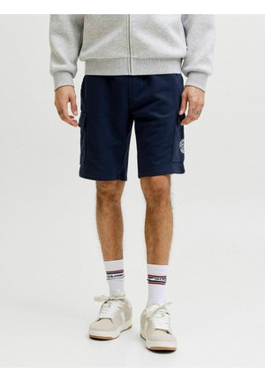 Uomo che indossa pantaloncini cargo blu navy, calzini bianchi con logo, sneakers beige e una felpa leggera grigia con cerniera, in piedi davanti a uno sfondo bianco neutro.
