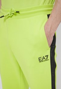 Neonově zelené tepláky s elastickým pasem, stahovací šňůrkou, boční kapsou a černým postranním pruhem s brandingem 'EA7 EMPORIO ARMANI'.