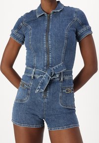 Denim jumpsuit i mörkblått med dragkedja fram, korta ärmar och bälte i midjan. Har två framfickor med guld detaljer.