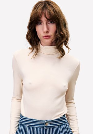 Vrouw met golvend bruin haar, gekleed in een beige coltrui en blauw-witte gestreepte broek, kijkt recht in de camera.