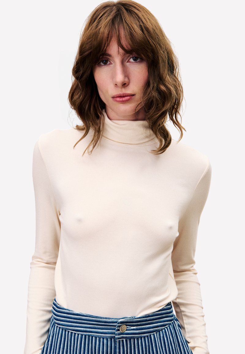 Frau mit welligem braunem Haar, die einen beigen Rollkragenpullover und eine blau-weiß gestreifte Hose trägt und direkt in die Kamera schaut.