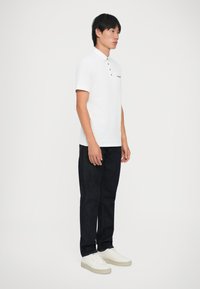 Hvid polo skjorte med tre knapper og lille logo, parret med mørke denim jeans og hvide sneakers, vist på en stående model.