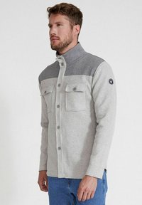 Hellgraue Knopfjacke mit einem dunkelgrauen oberen Bereich, flachen Taschen und einem hohen Kragen, gefertigt aus weichem Stoff, mit einem Logo am Ärmel.