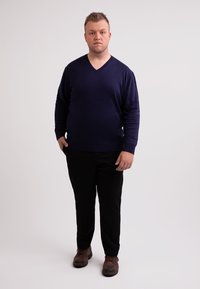Navy-blaues V-Ausschnitt-Pullover aus einem glatten Strickstoff, kombiniert mit schwarzen Hosen und braunen Schuhen. Einfaches, tailliertes Design mit langen Ärmeln.