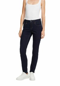 Žena v tmavě modrých úzkých džínách, bílém topu bez rukávů a bílých teniskách, stojící s uvolněnýma rukama podél těla na bílém pozadí.