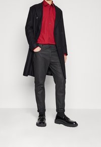 Schwarzer Mantel über einem roten Hemd mit Knöpfen, kombiniert mit grauer Hose und glänzenden schwarzen Schuhen. Verfügt über ein maßgeschneidertes Design mit Seitentaschen.