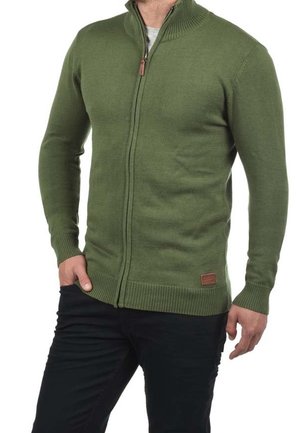 Homme portant un pull en maille vert à fermeture éclair intégrale avec poignets et ourlet côtelés, assorti à un pantalon noir, main dans la poche.