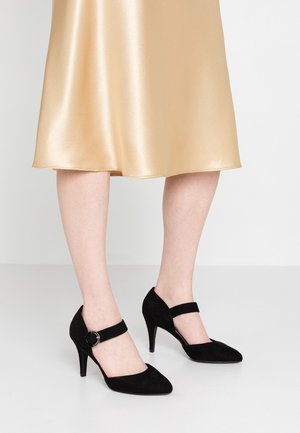 Legs wearing a beige satin midi skirt and black suede Mary Jane high heels with ankle straps, standing on a white floor.