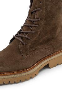 Botas de ante marrón con cordones, con detalles de costura y suela rugosa color marrón claro con profundas huellas, mostradas en un ángulo de cerca.