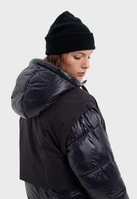 Chaqueta acolchada corta negra con secciones brillantes aislantes y capucha desmontable. Combinada con un gorro de punto negro con canalé.