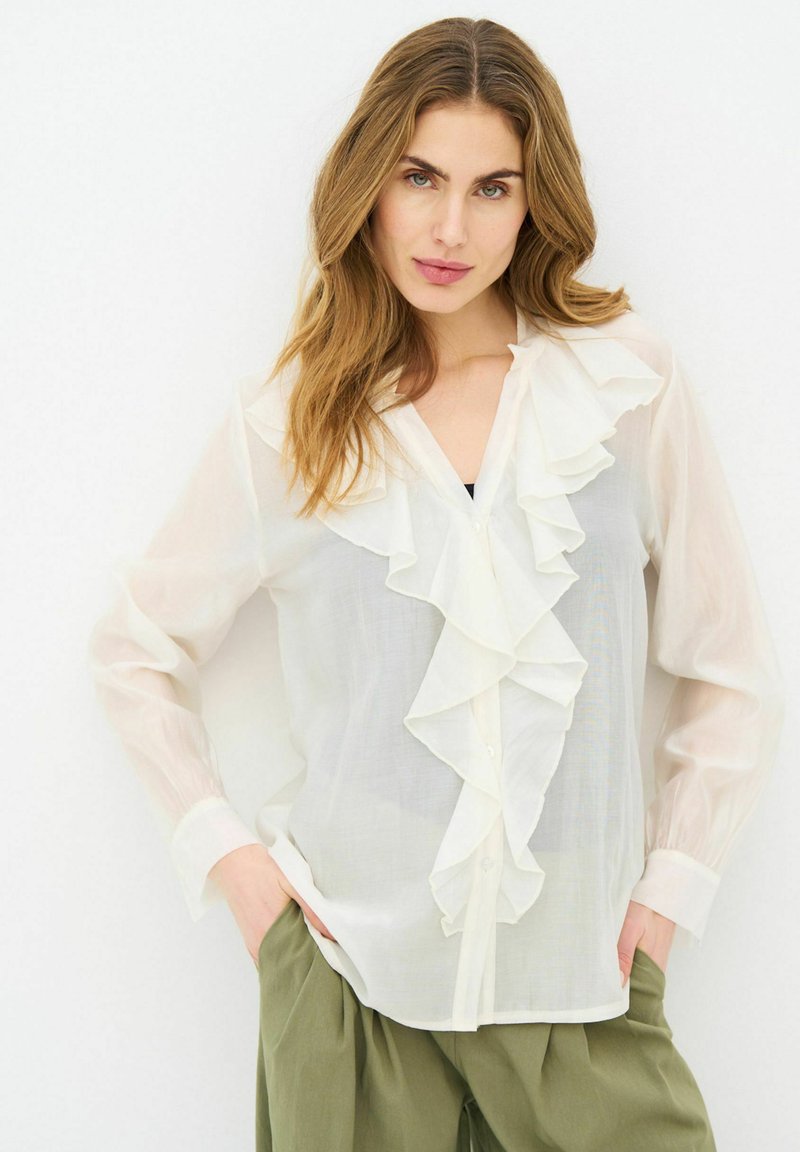 Jeune femme aux cheveux châtain clair portant un chemisier blanc transparent à volants et un pantalon vert olive, debout les mains dans les poches devant un fond blanc.
