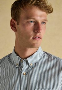 Koszula z krótkim rękawem, zapinana na guziki, z pionowymi niebiesko-białymi paskami, z kołnierzykiem typu spread oraz małymi okrągłymi guzikami o gładkiej fakturze.