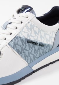 Baskets blanches avec des accents bleus, des superpositions texturées et un panneau à motif. Comprend un logo noir et un talon noir brillant. Semelle en caoutchouc.