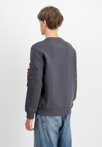 Szürke pulóver hosszú ujjaival, bordázott mandzsettákkal és zsebbel a bal ujján. Texturált anyag és kerek nyakkivágás.