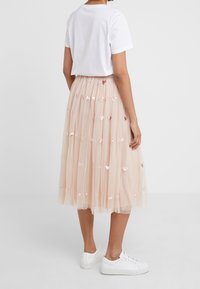 Vrouw van achteren gezien, gekleed in een wit T-shirt, een blush roze tulle midi rok met pailletten harten, en witte sneakers op een effen achtergrond.