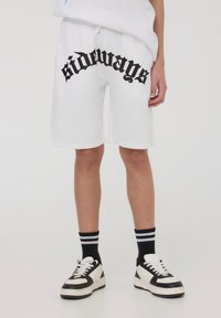 Shorts in cotone bianco con una grafica nera "di lato" sulla parte anteriore, caratterizzati da un fit comodo e una cintura elastica, abbinati a sneakers in bianco e nero.