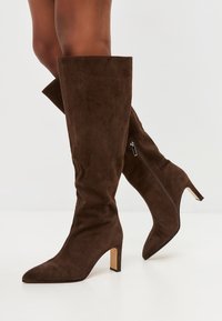 Stivali in suede marrone alti fino al ginocchio con punta affusolata e tacco a blocco di 5 cm. Presentano una zip laterale e una texture liscia.