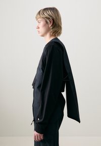 Crna jakna s zatvaračem i prevelikim stražnjim panelom. Materijal izgleda glatko s mat završnom obradom. Ima jednostavan, funkcionalan dizajn.