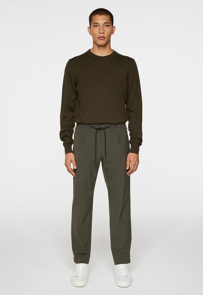 Temno zelena pletena pulover in usklajene tapered hlače z vrvico. Bele superge dopolnjujejo outfit, ki stoji pred enostavno podlago.