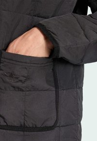 Zwarte pufferjack met een hand in de voorkzak, die de gequiltte textuur en subtiele logodetail langs de rand van de zak laat zien.