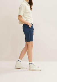 Jasno-beżowa koszula z kołnierzykiem i trzema brązowymi guzikami, w połączeniu z ciemnoniebieskimi szortami i białymi butami typu high-top z niebieskimi akcentami.