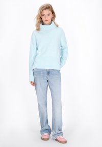 Gaiši zils, masīvs adīts turtlenecks džemperis ar ribotu apakšmalu, pārī ar gaišām džinsu biksēm un rozā sporta apaviem, pozēts uz vienkārša fona.