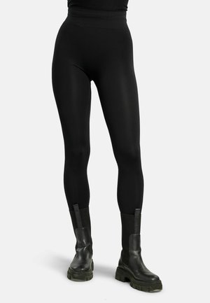 Person, die enge schwarze Leggings und klobige schwarze Leder-Stiefeletten trägt und vor einem schlichten weißen Hintergrund steht.