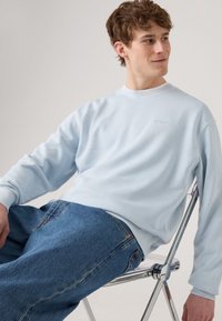 Jonge man met krullend haar die zijwaarts op een metalen stoel zit, gekleed in een lichtblauwe sweatshirt en blauwe jeans, kijkend naar rechts.