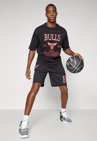 Sort basketball-tema outfit med en t-shirt med rød "BULLS" grafik og shorts med logo. Spilleren holder en basketball.