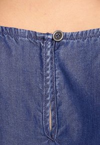 Blue denim top featuring a gathered neckline, a button closure, and a vertical slit detail at the back. The fabric is textured and durable.