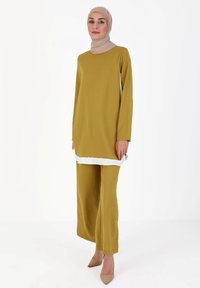 Mustard-colored tunic with white trim, long sleeves, and matching wide-leg pants. Smooth fabric and relaxed silhouette; paired with beige heels.