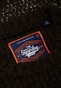 Tummanruskea neulepusero, jossa on sininen etiketti oransseilla yksityiskohdilla, teksti "Superdry Brand" ja lisäbrändäystietoja.