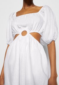 Valkoinen pellavamekko, jossa on neliömäinen pääntie, lyhyet puffihihat ja pyöreä leikkaus yksityiskohtana vyötäröllä. Teksturoitu, virtaava kangas.