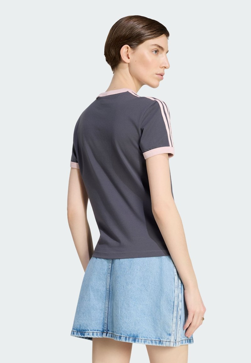 Kvinde med kort brunt hår iført en mørkegrå t-shirt med pink kant og striber, kombineret med et lyseblåt denimskørt, set fra siden.