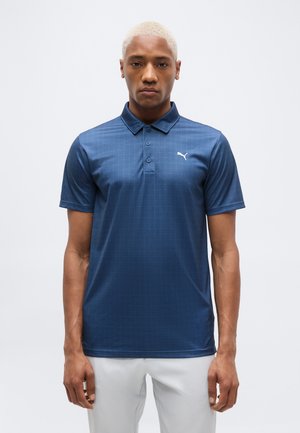 Ung mand med kort, bleget hår iført en mørkeblå Puma polo og hvide bukser, stående foran en ensfarvet lys baggrund.